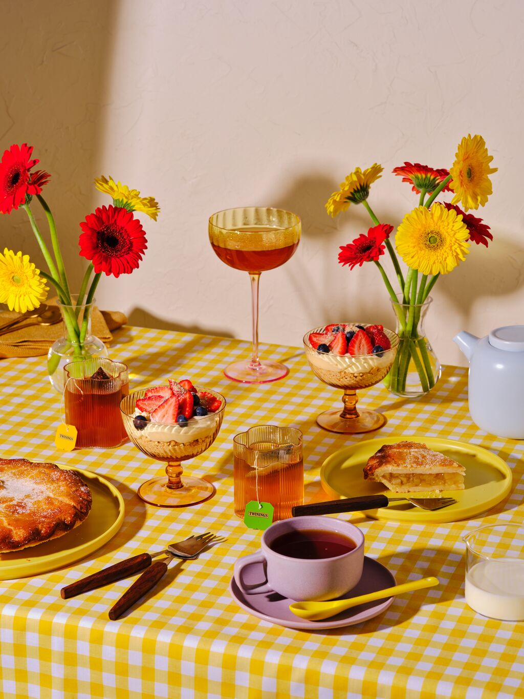 Table setting with a yellow checkered tablecloth, flowers, and various dishes including a pie and tea.