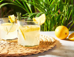 Two glasses of lemonade with lemon slices and a sprig of rosemary on a woven mat with lemons and greenery in the background.