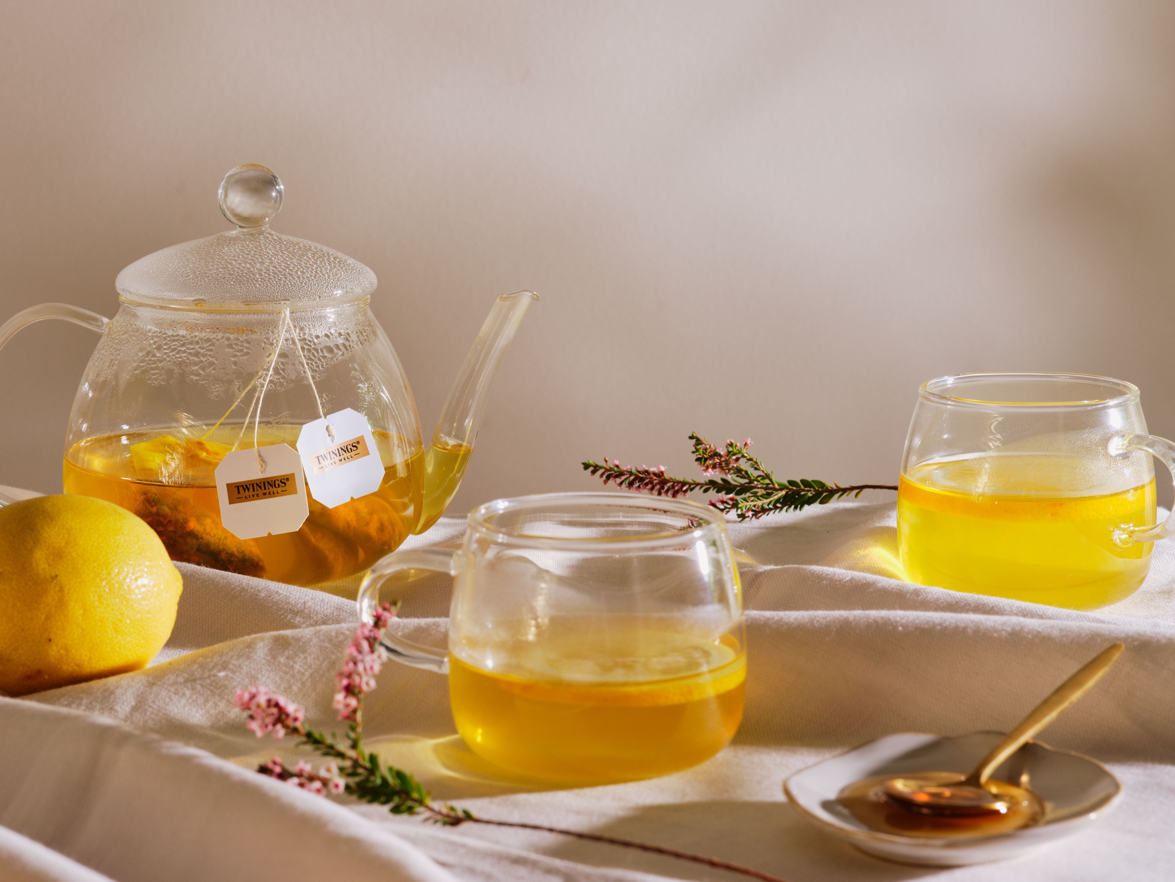 Clear glass teapot and cups with lemon slices and flowers on a white cloth.