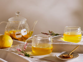 Clear glass teapot and cups with lemon slices and flowers on a white cloth.