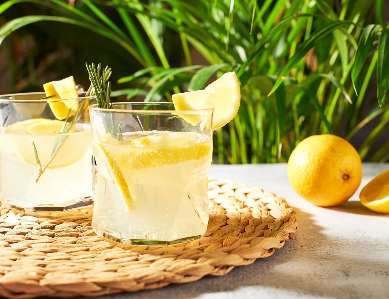 Two glasses of lemonade with lemon slices and a sprig of rosemary on a woven mat with lemons and greenery in the background.