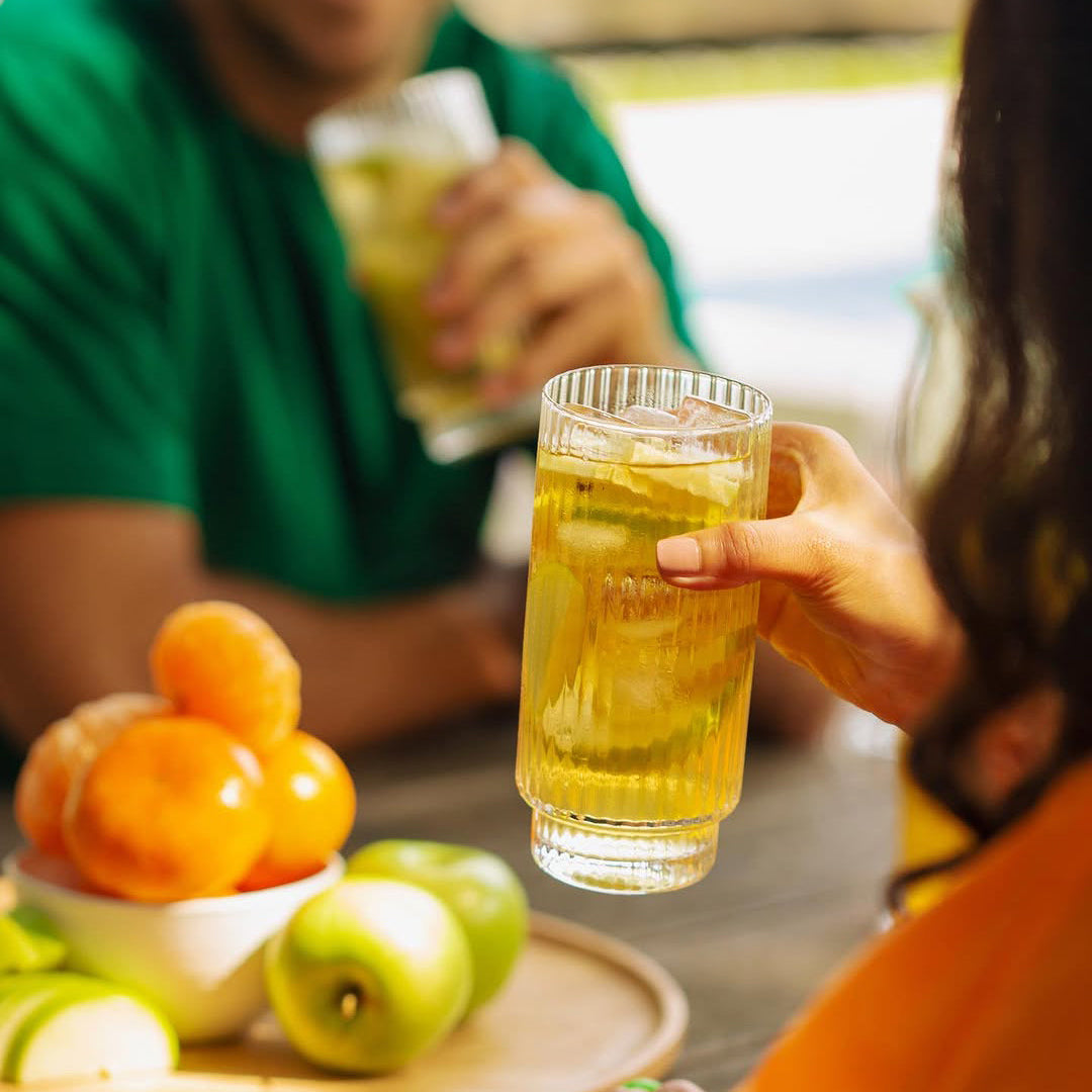 Two people holding glasses of iced tea with a bowl of fruit on a table.